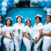 Group of six nurses in white uniforms holding roses, standing in front of a blue backdrop with white and blue balloons above.